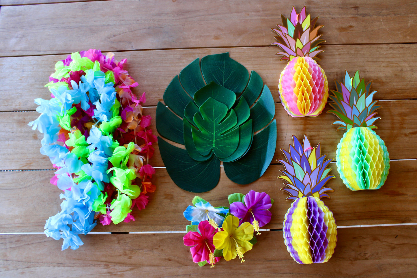 Présentation des accessoires présents dans la box.
Les colliers de fleurs artificielles, les monstera artificielles, les fleurs tropicales artificielles et les trois ananas en papier sont posés sur la table, sans emballage