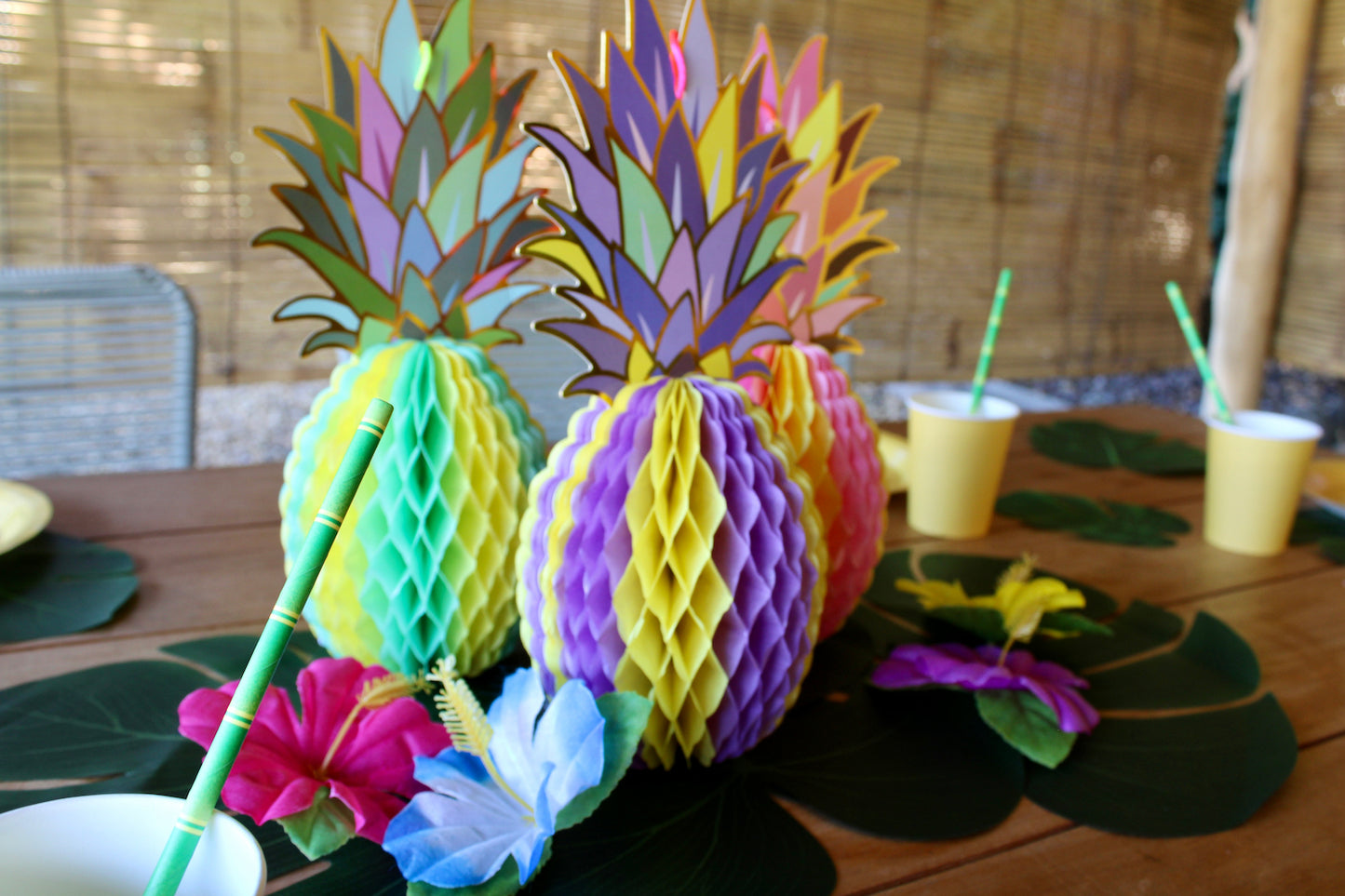 Au centre, il y a trois grandes monstera posées en quinconce, avec au dessus trois ananas en papier pliables. Un est jaune et bleu turquoise, un autre violet et jaune, et enfin le troisième est rose et jaune. Autour de ces ananas il y a quatre fleurs tropicales artificielles de différentes couleurs.