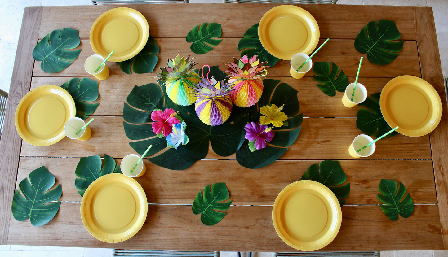 Table en bois avec des monstera artificielles de différentes tailles un peu partout. Au centre, il y a trois grandes monstera posées en quinconce, avec au dessus trois ananas en papier pliables. Un est jaune et bleu turquoise, un autre violet et jaune, et enfin le troisième est rose et jaune. Autour de ces ananas il y a quatre fleurs tropicales artificielles de différentes couleurs.
La table est dressée avec six assiettes, six gobelets, six serviettes, en papier et en carton et jaune citron.