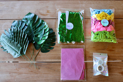 Présentation des accessoires présents dans la box.
Tout à gauche, les six feuilles de monstera avec les pics en fer, à droite les deux rideaux à franges verts emballés dans un papier transparent avec un sticker holographique où il est écrit "DAVEIS". En dessous le ballon flamant rose dégonflé et emballé. À droite un petit pochon holographique avec deux rouleaux à l'intérieur : un rouleau de ruban perforé et un rouleau de points de colle. Au-dessus, les ballons dégonflés emballés  comme les rideaux.