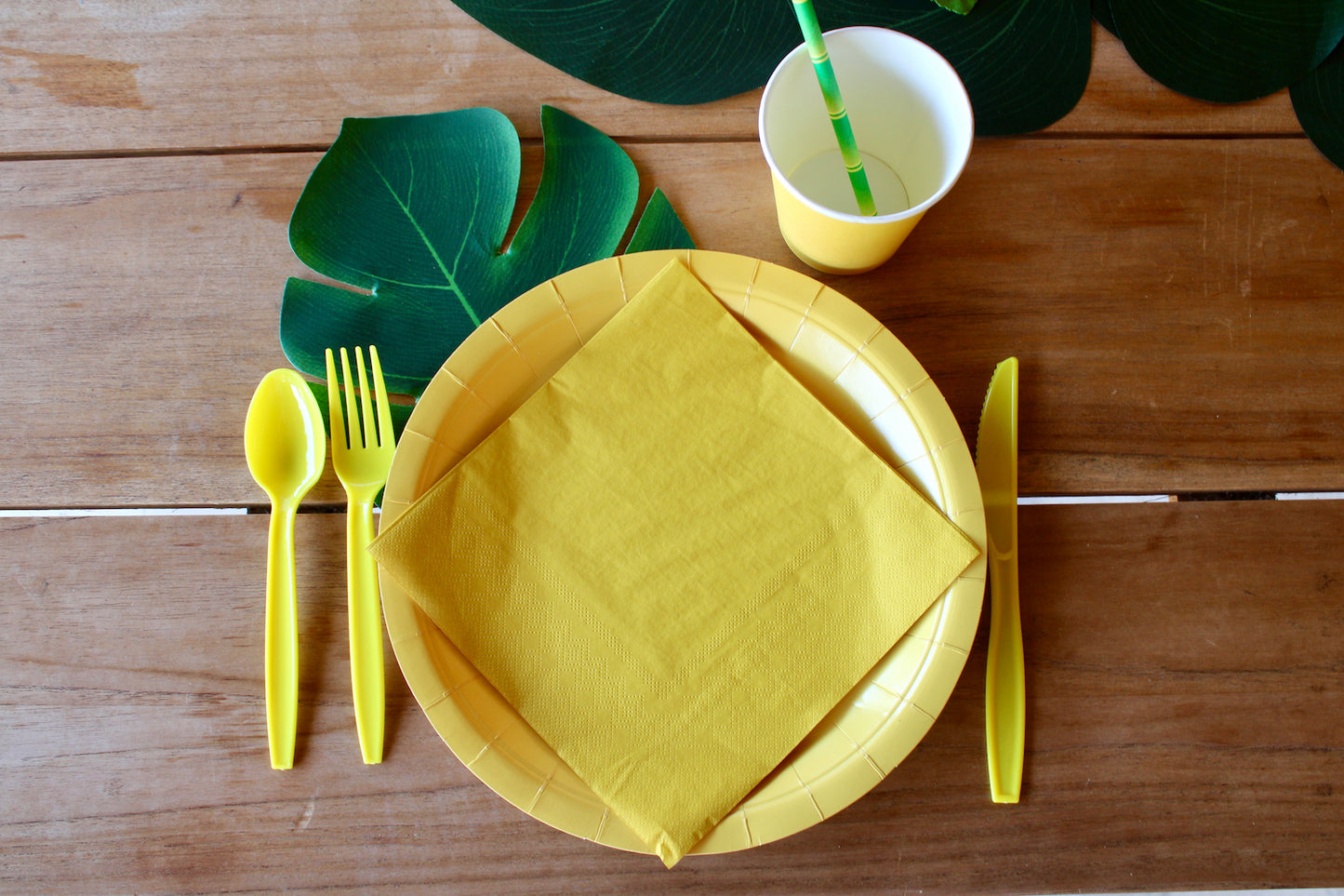 Zoom sur le dressage par personne : Une assiette en carton jaune citron, une serviette en papier jaune citron posée dessus. Un gobelet en carton jaune citron avec une paille en carton imprimée bambou à l'intérieur. Et enfin, des couverts en plastique jaune citron. La cuillère et la fourchette à gauche et le couteau à droite.